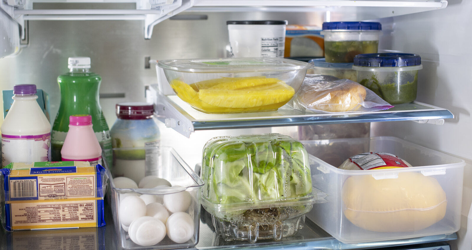 A real household refrigerator containing sealed leftovers, dairy, eggs, produce, bread, and cheese